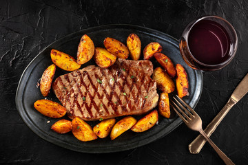 Grilled beef steak with baked potato wedges and red wine, shot from above on a black background