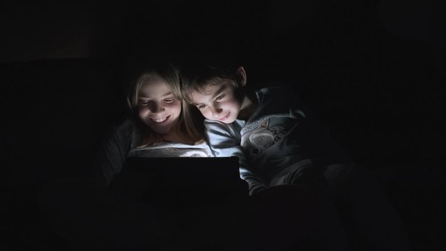 Video of cute small boy and girl lying under the blanket at night and watching video on tablet computer.