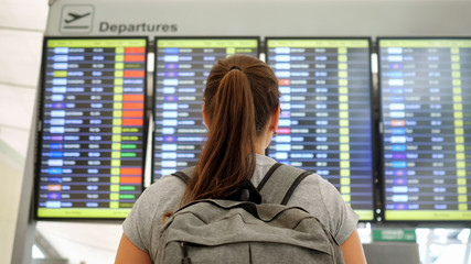 brunette with ponytail and grey backpack wearing t-shirt looks at departures schedule in airport lounge backside view