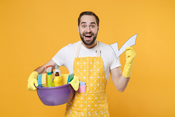 Excited young man househusband in apron rubber gloves hold basin detergent bottles washing...