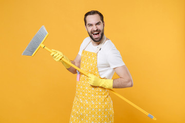 Laughing cheerful young man househusband in apron rubber gloves hold in hands broom while doing...