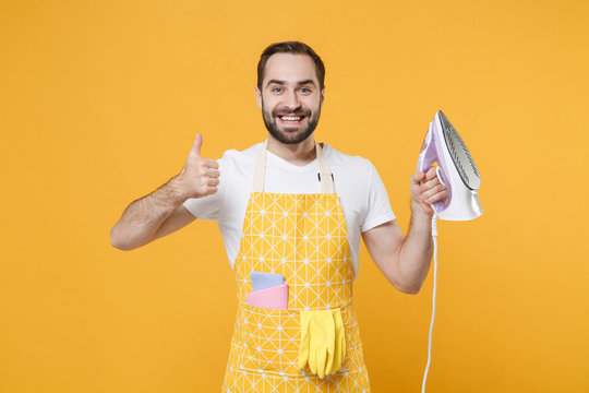 Smiling Attractive Young Man Househusband In Apron Hold In Hands Iron While Doing Housework Isolated On Yellow Background Studio Portrait. Housekeeping Concept. Mock Up Copy Space. Showing Thumb Up.