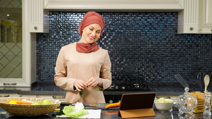 smiling lady in hijab talks with relatives on Skype using modern laptop and cooking supper in kitchen close view