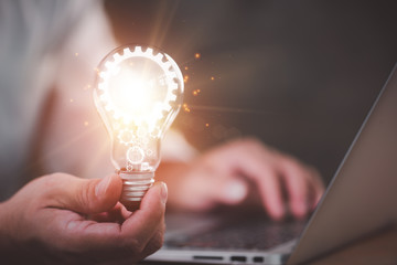 The hand of a man with a light bulb And there is a gear icon in it And he is using a notebook...