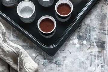 Chocolate dough for muffins in paper molds on a black baking sheet. On a gray wooden messy background. Cooking at home.