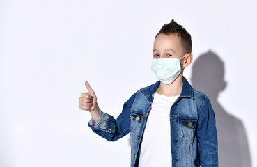 Teenage boy in a blue denim jacket, t-shirt. He is in a medical mask, showing thumb up, posing isolated on a white studio background.