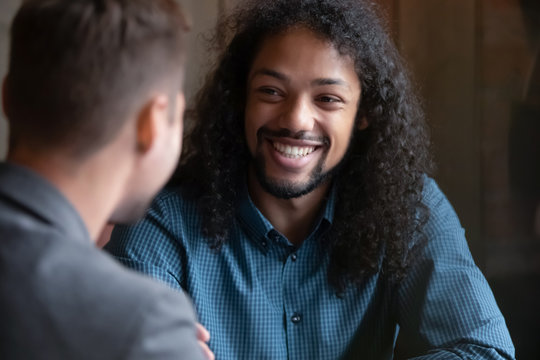 Smiling biracial young man with long hair sit in cafe talking with male friend, happy overjoyed millennial multiethnic guy meeting with colleague coworker, have fun chilling in coffeeshop together - Powered by Adobe