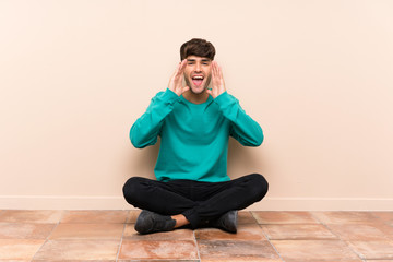 Young handsome man sitting on the floor shouting with mouth wide open