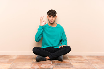 Young handsome man sitting on the floor showing an ok sign with fingers