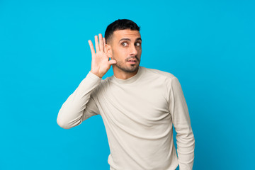 Young man over isolated blue background listening to something by putting hand on the ear