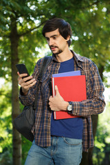 Young man looking at screen of smartphone walking in a park with backpack and books for education