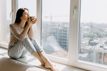 Young woman sitting on a windowsill by the window with a city view, drinking coffee or tea in the...