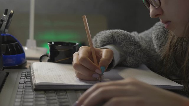 Close-up, teenage girl doing homework on the distance learning front of a laptop. Girl with a pencil writing in a notebook.