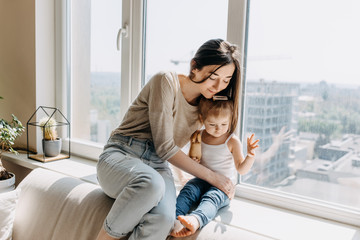 Mother and daughter staying home, sitting on windowsill, hugging. 