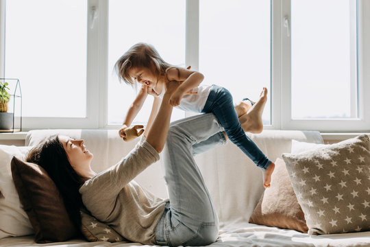 Mother and little daughter playing together, lying on the sofa by the window.
