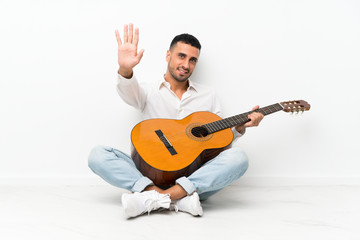 Obraz premium Young man sitting on the floor with guitar saluting with hand with happy expression