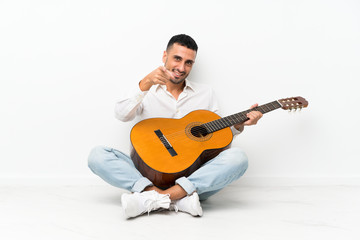 Young man sitting on the floor with guitar points finger at you with a confident expression