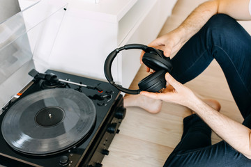 Young hipster man, sitting on the floor, listening to music on a vinyl player, looking to a vinyl...