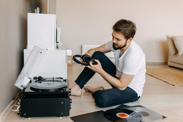 Young hipster man with beard, sitting on the floor, listening to music on a vinyl player, looking...