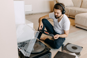Young hipster man with beard, sitting on the floor, listening to music on a vinyl player, using...