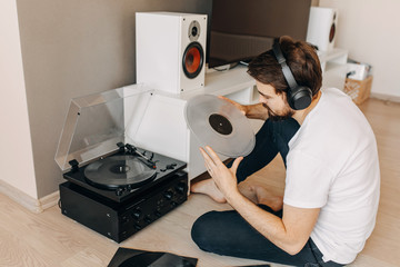 Young hipster man with beard, sitting on the floor, listening to music on a vinyl player, using...