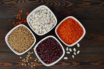 Set of various dried legumes in ceramic bowls: red lentils, green lentils, red beans, white beans on a dark wooden background, top view.