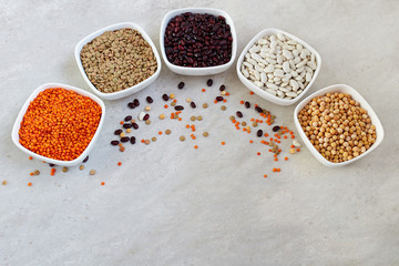 A set of different dried legumes in ceramic bowls: red lentils, green lentils, peas, red beans, white beans on grey background