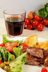 pieces of grilled steak next to potatoes grilled next to lettuce with tomatoes and olives and a drink in a glass