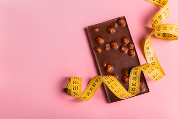 Diet concept with chocolate bar and measuring tape for weight loss on the pink background. Top view...