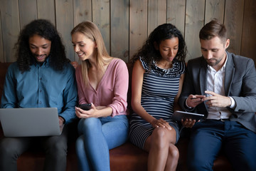Diverse young people sit on couch in coffeeshop have fun talking using modern gadgets, multiracial friends or colleagues relax rest on sofa in cafe addicted to modern technological electronic devices