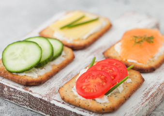 Obraz na płótnie Canvas Various healthy crackers with salmon and cheese, tomato and cucumber on wooden chopping board on light kitchen table background. Macro