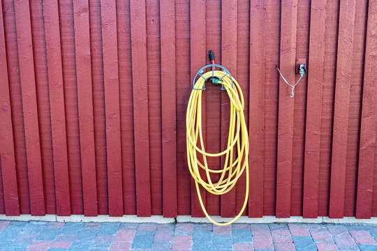 Yellow rubber watering tube for plants watering hangs on red wooden wall of traditional Swedish garden shed close to wall mounted water tap. Decorative stones, sunny Scandinavian summer