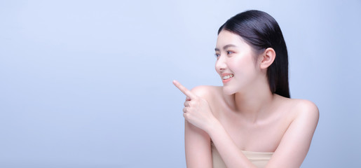 Beauty spa woman with perfect skin portrait. Beautiful asian spa girl smiling and showing empty copy space on index finger for text. Proposing product. Gestures for advertisement. Blue background.
