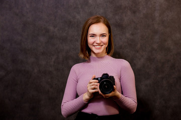 a young beautiful girl photographer holds her camera, looks at the camera and smiles,she takes pictures, she loves the camera and her work. On dark background