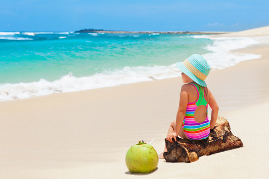 Little baby girl in hat sit on white sand beach. Child drink young coconut, look at ocean and enjoying. Kids travel lifestyle. Family summer holiday. Activity on tropical Hawaii and Caribbean islands.