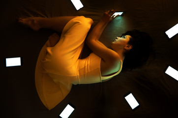 Cute girl in orange dress lying in the dark on her bed surrounding with cellphones conception mean...