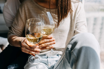 Close-up of a young couple of man and woman in casual clothes, sitting on windowsill in day light hugging and drinking wine.