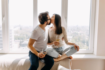 Young couple of man and woman in casual clothes, sitting on windowsill in day light hugging, kissing and drinking wine.