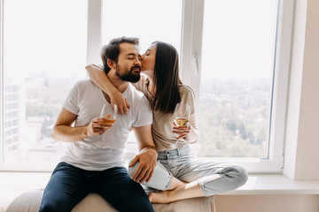 Young couple of man and woman in casual clothes, sitting on windowsill in day light hugging, kissing on cheek and drinking wine.
