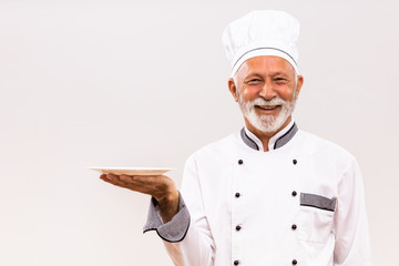 Portrait of senior chef with empty plate on gray background.
