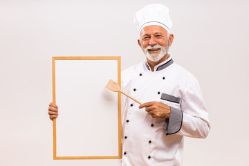 Portrait of senior chef showing with wooden spoon whiteboard on gray background.