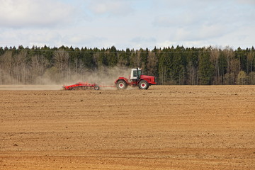 Obraz premium Red big wheeled tractor with large plough ploughs the land in a field on forest background at spring day, rural farm landscape