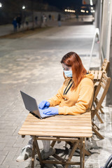 A woman in a medical mask and gloves remotely works late at night outdoors. Ocher-colored hoodie girl with laptop on the street during quarantine. Coronavirus.