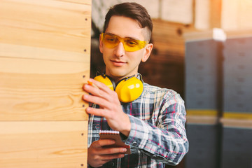 Young man warehouse manager in protective glasses, headphones counting wood manufacturing material and smiling. Male sawmill worker inspecting wood production factory inventory. Small business owner