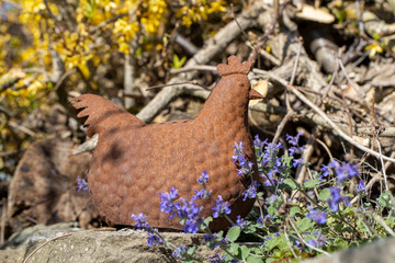 Ein rostiges Blechhuhn in der Seitenansicht im Vordergrund blaue Blüten und im Hintergrund gelbe Blüten, Blechhuhn, Metall,