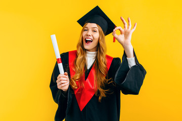 graduate shows the OK gesture with a diploma in her hands on a yellow background. Concept of the graduation ceremony