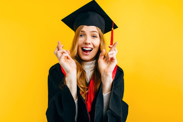 Beautiful graduate girl praying for success with crossed fingers on a yellow background