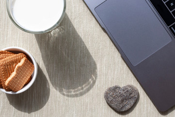 A glass of milk and a saucer with cookies and small stone with sharp sunny shadows and a fragment of a laptop