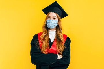 beautiful graduate woman wearing a medical protective mask on an isolated yellow background. Graduation, quarantine, coronavirus
