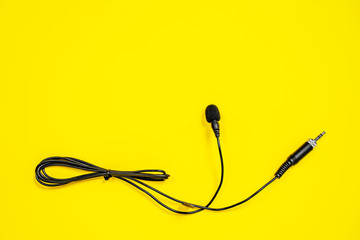 Lapel microphone with a bundled cord on yellow background, top view. Essential vlogging and journalism tools. Piece of audio equipment designed to record sounds and speech.